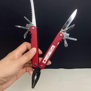 Puede incluir: Una herramienta multifunción roja con alicates y varios accesorios desplegables, incluyendo una hoja, una sierra y una regla. El mango tiene una etiqueta blanca con una cruz roja y el texto METRIC CONVERSIONS, mostrando varias unidades.