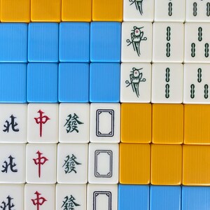 Puede incluir: Vista a&eacute;rea de fichas de Mahjong dispuestas en una cuadr&iacute;cula. Las fichas muestran varios s&iacute;mbolos, caracteres y colores, incluyendo azul, amarillo y blanco. Algunas fichas presentan caracteres chinos.