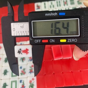 Puede incluir: Una ficha de Mahjong blanca y roja se mide con un calibrador digital, mostrando un ancho de 1,64 cm. La ficha presenta caracteres chinos y est&aacute; rodeada de otras fichas con detalles rojos y verdes. El calibrador muestra "mm/inch".