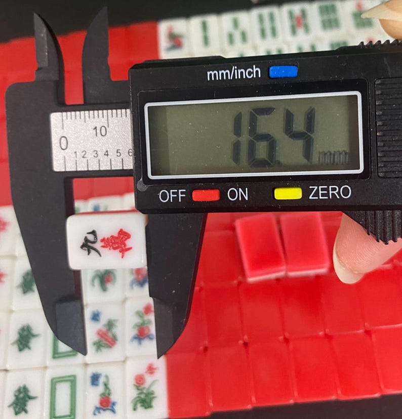 Puede incluir: Una ficha de Mahjong blanca y roja se mide con un calibrador digital, mostrando un ancho de 1,64 cm. La ficha presenta caracteres chinos y est&aacute; rodeada de otras fichas con detalles rojos y verdes. El calibrador muestra "mm/inch".