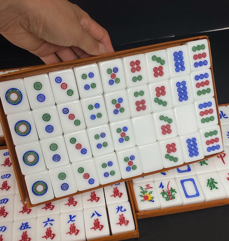 May include: A set of white Mahjong tiles with colourful markings, including circles and symbols in red, green, and blue. The tiles are arranged in a wooden box, with some tiles visible in a second box below.