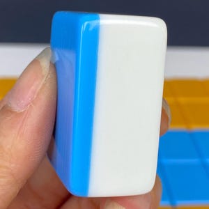 Puede incluir: Una ficha de juego rectangular de dos tonos. Un lado es blanco y el otro azul vibrante. La ficha se sostiene en una mano, mostrando su superficie lisa y brillante. Probablemente parte de un juego de mesa.