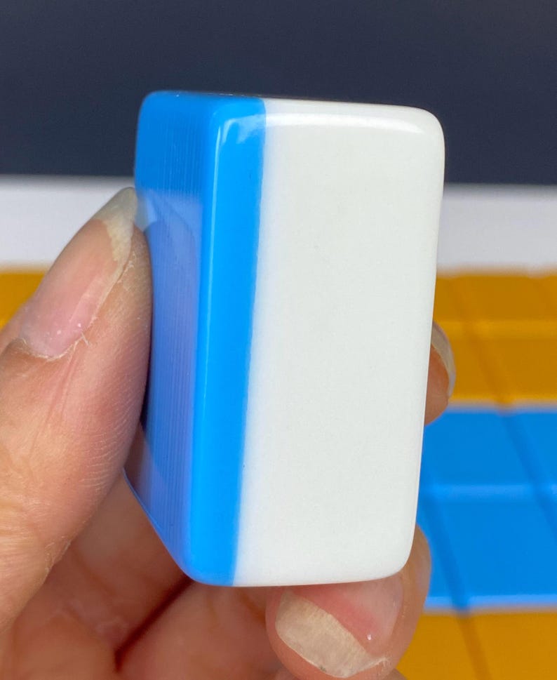 Puede incluir: Una ficha de juego rectangular de dos tonos. Un lado es blanco y el otro azul vibrante. La ficha se sostiene en una mano, mostrando su superficie lisa y brillante. Probablemente parte de un juego de mesa.