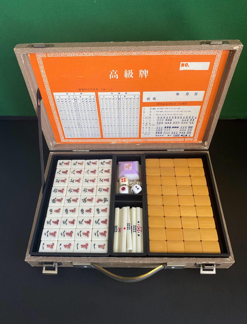 May include: Open wooden Mahjong set with tiles, dice, and racks. The set includes tiles with Chinese characters and symbols, dice, and wooden racks. The case is open, revealing the game pieces. The interior is lined with orange paper.
