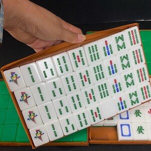 May include: A set of white Mahjong tiles with green, red, and blue markings, presented in a brown box. Some tiles feature a colorful bird design. The game set is partially open, revealing the tiles.
