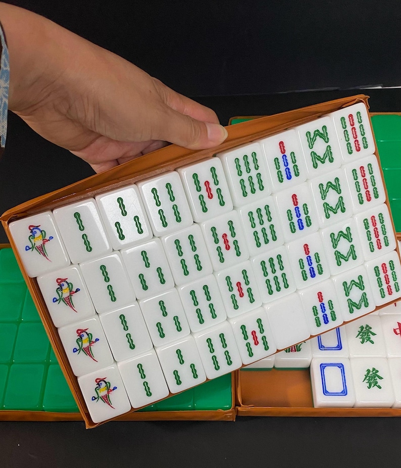 May include: A set of white Mahjong tiles with green, red, and blue markings, presented in a brown box. Some tiles feature a colorful bird design. The game set is partially open, revealing the tiles.