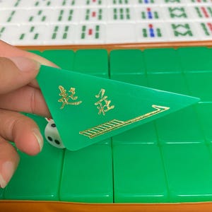 May include: A green triangular Mahjong tile with gold Chinese characters and an arrow design. The tile is held over a set of green Mahjong tiles and a white die.