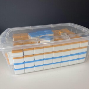 Puede incluir: Un juego de Mahjong en un recipiente de pl&aacute;stico transparente. Las fichas son blancas con detalles azules y naranjas. Se ven un par de dados. El recipiente tiene una tapa transparente.
