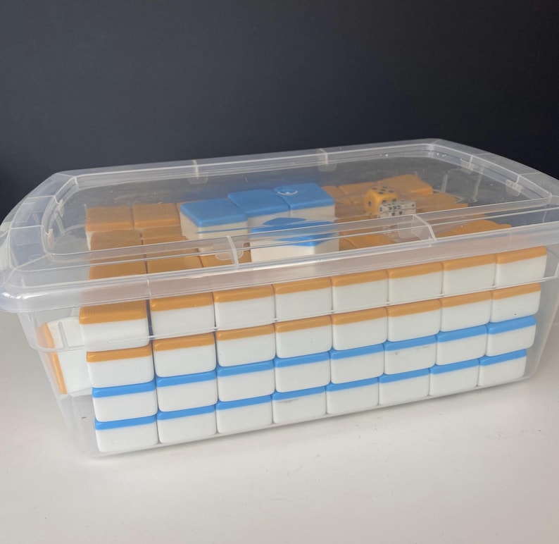 Puede incluir: Un juego de Mahjong en un recipiente de pl&aacute;stico transparente. Las fichas son blancas con detalles azules y naranjas. Se ven un par de dados. El recipiente tiene una tapa transparente.