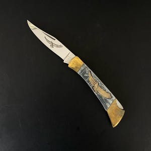 May include: A folding pocket knife with a silver blade and a handle featuring an engraved eagle design. The handle has gold-colored accents and a silver and gray background. The blade has an engraved eagle. The word "KEEORS" is printed on the blade.