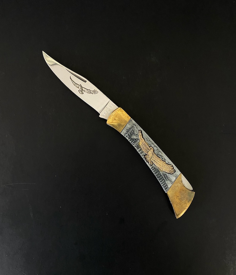 May include: A folding pocket knife with a silver blade and a handle featuring an engraved eagle design. The handle has gold-colored accents and a silver and gray background. The blade has an engraved eagle. The word "KEEORS" is printed on the blade.