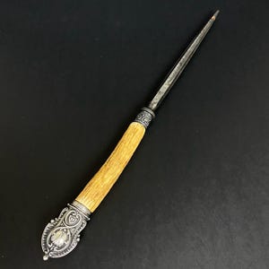 Könnte beinhalten: Ein antiker Brieföffner mit hellbraunem Griff und einem dekorativen Ende aus silberfarbenem Metall. Die Klinge ist lang und schlank, mit einer Spitze. Der Griff besteht aus einem hellbraunen Material, möglicherweise Knochen oder Geweih.