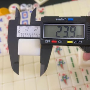 May include: A black digital caliper measuring a white tile. The display reads 23.9 mm. The tile is part of a Mahjong set, with other tiles visible. The caliper has mm/inch markings. The tile's measurement is approximately 0.94 inches.