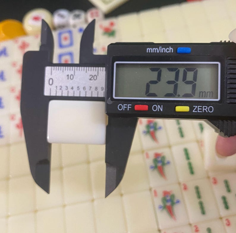 May include: A black digital caliper measuring a white tile. The display reads 23.9 mm. The tile is part of a Mahjong set, with other tiles visible. The caliper has mm/inch markings. The tile's measurement is approximately 0.94 inches.