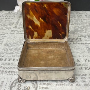 Op de afbeelding: Een zilveren en schildpadschatulle met een fluwelen interieur. De doos heeft vier pootjes en is versierd met een bloemmotief.