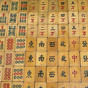 May include: A collection of vintage Mahjong tiles, featuring a variety of symbols and characters in red and black ink. The tiles are beige with intricate designs, including circles, bamboo, and Chinese characters. The game pieces are arranged in a grid pattern.