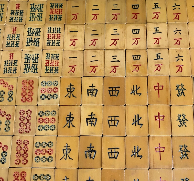 May include: A collection of vintage Mahjong tiles, featuring a variety of symbols and characters in red and black ink. The tiles are beige with intricate designs, including circles, bamboo, and Chinese characters. The game pieces are arranged in a grid pattern.