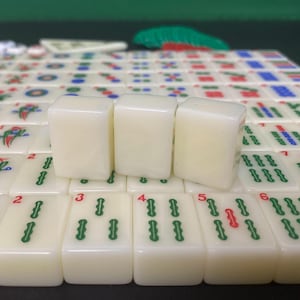 May include: A close-up of ivory-coloured Mahjong tiles with green and red markings. Three tiles are in focus, with others in the background. The tiles display numbers and symbols, set on a green surface.
