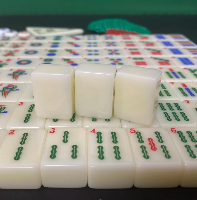 May include: A close-up of ivory-coloured Mahjong tiles with green and red markings. Three tiles are in focus, with others in the background. The tiles display numbers and symbols, set on a green surface.