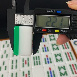 May include: A white and green Mahjong tile is measured with a digital caliper. The tile is 2.21 cm wide. Other Mahjong tiles are visible in the background.