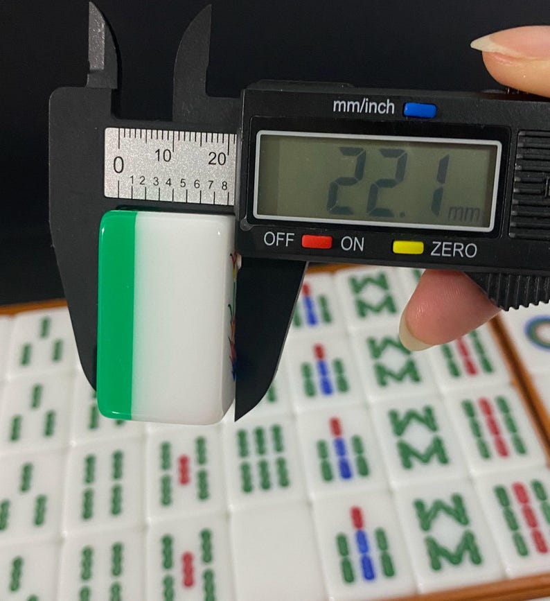 May include: A white and green Mahjong tile is measured with a digital caliper. The tile is 2.21 cm wide. Other Mahjong tiles are visible in the background.