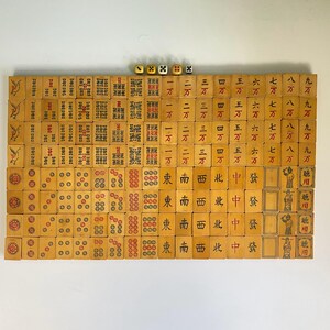 May include: A complete set of vintage Mahjong tiles arranged on a white surface. The tiles are beige with red, black, and green markings. Four dice are visible at the top. The game set includes tiles with Chinese characters and symbols.