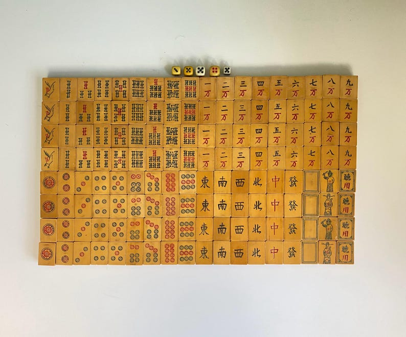 May include: A complete set of vintage Mahjong tiles arranged on a white surface. The tiles are beige with red, black, and green markings. Four dice are visible at the top. The game set includes tiles with Chinese characters and symbols.
