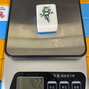Puede incluir: Una ficha de Mahjong blanca con un gr&aacute;fico verde y rojo, colocada sobre una b&aacute;scula digital. La b&aacute;scula muestra "42.2" y tiene caracteres chinos. La ficha tiene un borde azul. Otras fichas de Mahjong son visibles en el fondo.