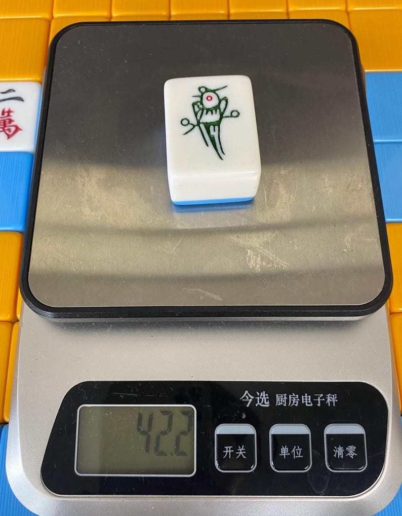 Puede incluir: Una ficha de Mahjong blanca con un gr&aacute;fico verde y rojo, colocada sobre una b&aacute;scula digital. La b&aacute;scula muestra "42.2" y tiene caracteres chinos. La ficha tiene un borde azul. Otras fichas de Mahjong son visibles en el fondo.