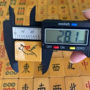 May include: A vintage Mahjong tile, measuring approximately 1.1 inches (28.1 mm), featuring a bird design in black and red. The tile is held by a digital caliper, with other tiles visible in the background.