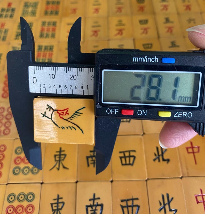 May include: A vintage Mahjong tile, measuring approximately 1.1 inches (28.1 mm), featuring a bird design in black and red. The tile is held by a digital caliper, with other tiles visible in the background.