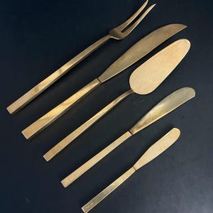 May include: A collection of six gold-coloured serving utensils, including a fork, knives, and a cake server, arranged diagonally on a dark surface. The utensils have a sleek, modern design, ideal for serving food.