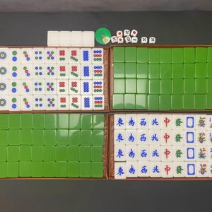 Puede incluir: Un juego de Mahjong con cuatro cajas rectangulares que contienen fichas blancas con patrones circulares y florales en azul, verde y rojo, fichas de color verde sólido y fichas blancas con caracteres chinos azules, dados pequeños y una ficha verde.