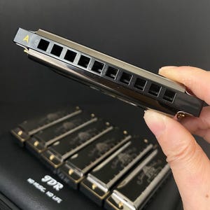 May include: A black harmonica with silver accents is held in a hand, with the letter "A" visible. Below, several harmonicas rest on a black case. The harmonicas have a black finish with gold-colored details and decorative designs.