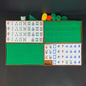 Puede incluir: Un juego de Mahjong completo con fichas blancas con varios símbolos y caracteres, dispuesto en una caja marrón con compartimentos verdes. Incluye fichas y dados de colores. Un juego de mesa clásico.