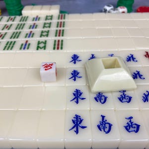 May include: A close-up of a Mahjong game set. The ivory-colored tiles have blue Chinese characters. A white die with red markings and a beige tile holder are also visible. Green and red game pieces are in the background.