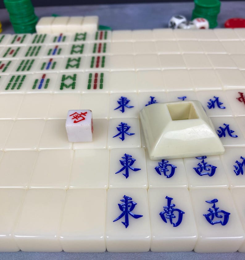 May include: A close-up of a Mahjong game set. The ivory-colored tiles have blue Chinese characters. A white die with red markings and a beige tile holder are also visible. Green and red game pieces are in the background.