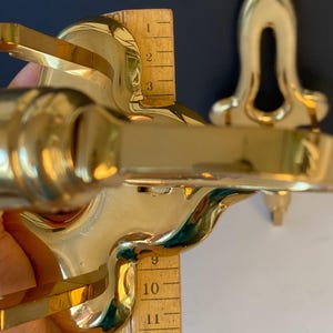 May include: Close-up of a shiny, gold-colored metal object with curved edges and a decorative design. A wooden ruler is placed next to the object, measuring approximately 12 inches. The object appears to be a decorative fixture.