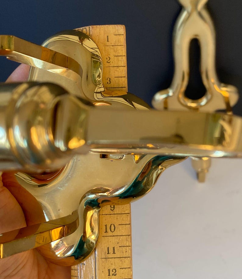 May include: Close-up of a shiny, gold-colored metal object with curved edges and a decorative design. A wooden ruler is placed next to the object, measuring approximately 12 inches. The object appears to be a decorative fixture.