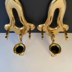 May include: Two ornate, gold-colored wall sconces. Each sconce features a curved, open design with a small, bell-shaped cup at the bottom. The sconces are made of a shiny, metallic material and are designed to hold candles or small objects.