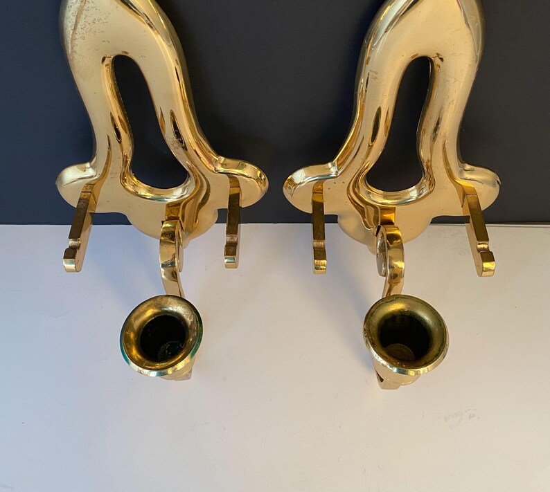 May include: Two ornate, gold-colored wall sconces. Each sconce features a curved, open design with a small, bell-shaped cup at the bottom. The sconces are made of a shiny, metallic material and are designed to hold candles or small objects.