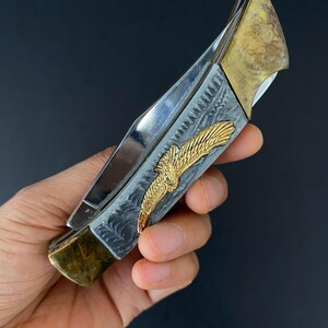 May include: A silver and gold folding knife with an eagle emblem. The knife features a polished steel blade, a handle with an engraved design, and gold-colored accents. The eagle emblem is in gold. The knife is held against a dark background.