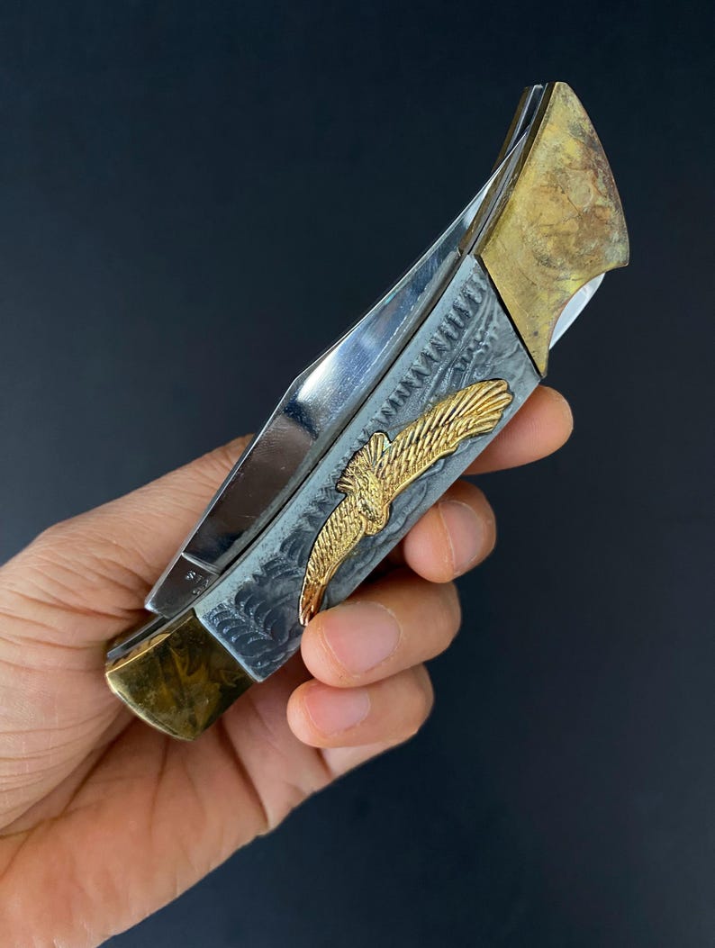 May include: A silver and gold folding knife with an eagle emblem. The knife features a polished steel blade, a handle with an engraved design, and gold-colored accents. The eagle emblem is in gold. The knife is held against a dark background.