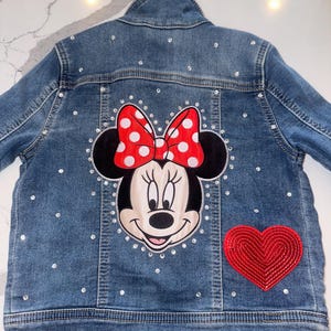May include: A blue denim jacket with a red and white Minnie Mouse applique on the back. The jacket is decorated with rhinestones and has a red heart applique on the bottom right side.