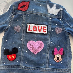 May include: A blue denim jacket with various patches, including a red lip, a white crown, a pink heart, a Mickey Mouse head, a Minnie Mouse head, and a white patch that says "LOVE".