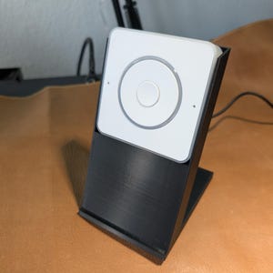以下が含まれることがあります： 前面に丸いボタンが付いた白いスマートスピーカーが、黒いスタンドの上に置かれています。スピーカーは黒いケーブルで電源に接続されています。