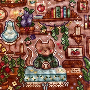 May include: Cross-stitch art featuring a cozy scene with a brown cat character in a green sweater, holding a cup. The scene includes bookshelves, candles, and floral accents, all in a warm, inviting color palette. The artwork is detailed and intricate.
