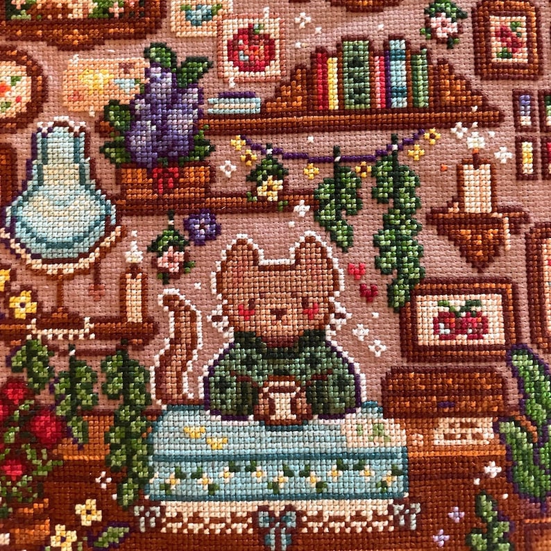 May include: Cross-stitch art featuring a cozy scene with a brown cat character in a green sweater, holding a cup. The scene includes bookshelves, candles, and floral accents, all in a warm, inviting color palette. The artwork is detailed and intricate.