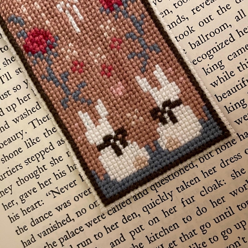 May include: A cross-stitch bookmark featuring two white bunnies with black bows, standing in front of a pink and brown background. The bookmark is framed with a dark blue border.