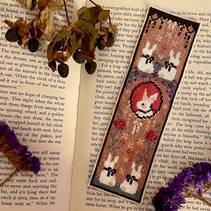 May include: A brown bookmark with a cross-stitch design featuring three white rabbits with black eyes and noses. The rabbits are surrounded by red, blue, and green flowers. The bookmark has the text "The Little Rabbit" in white lettering.